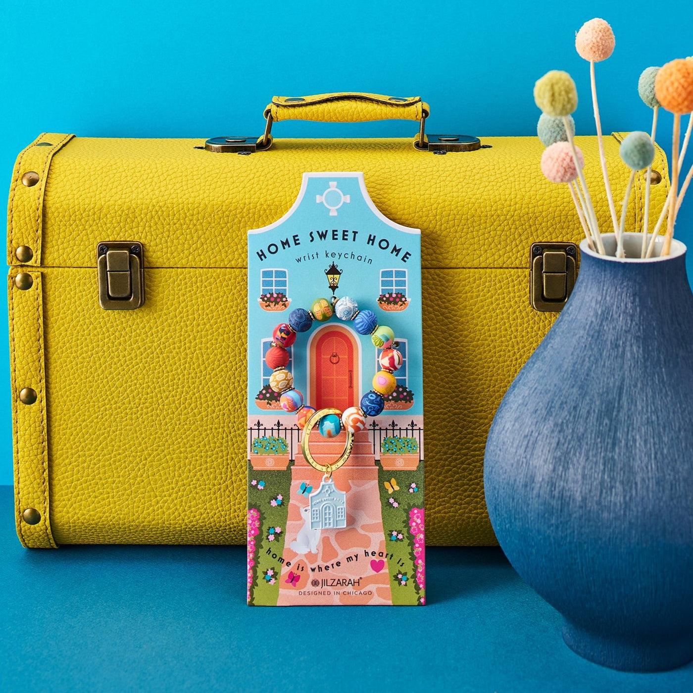 Happy Place Home Sweet Home wrist keychain propped against a yellow suitcase on a turquoise backdrop