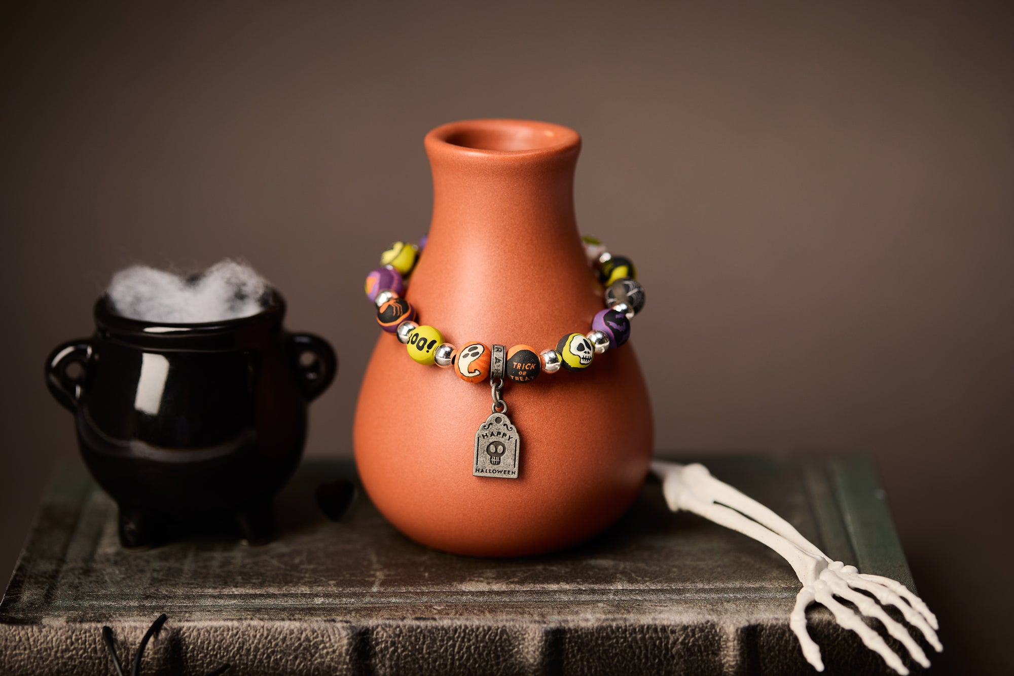 JILZARAH Halloween bracelet on an orange vase beside a smoking cauldron and a skeleton hand
